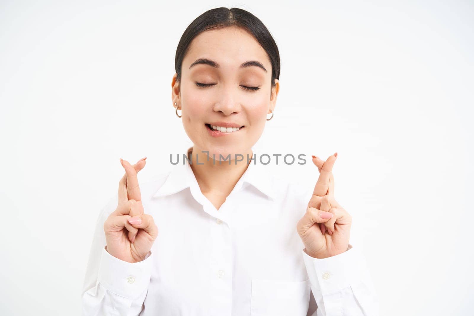 Close up portrait of hopeful businesswoman, cross fingers for good luck, waits for something, makes wish, smiles and anticipates, white studio background.