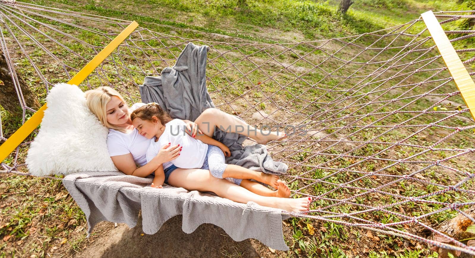 Wide view of a young mother and daughter relaxing together and smiling sitting in a hammock, hugging and lounging during a sunny summer day in a holiday home garden with grass and trees, lifestyle by Andelov13