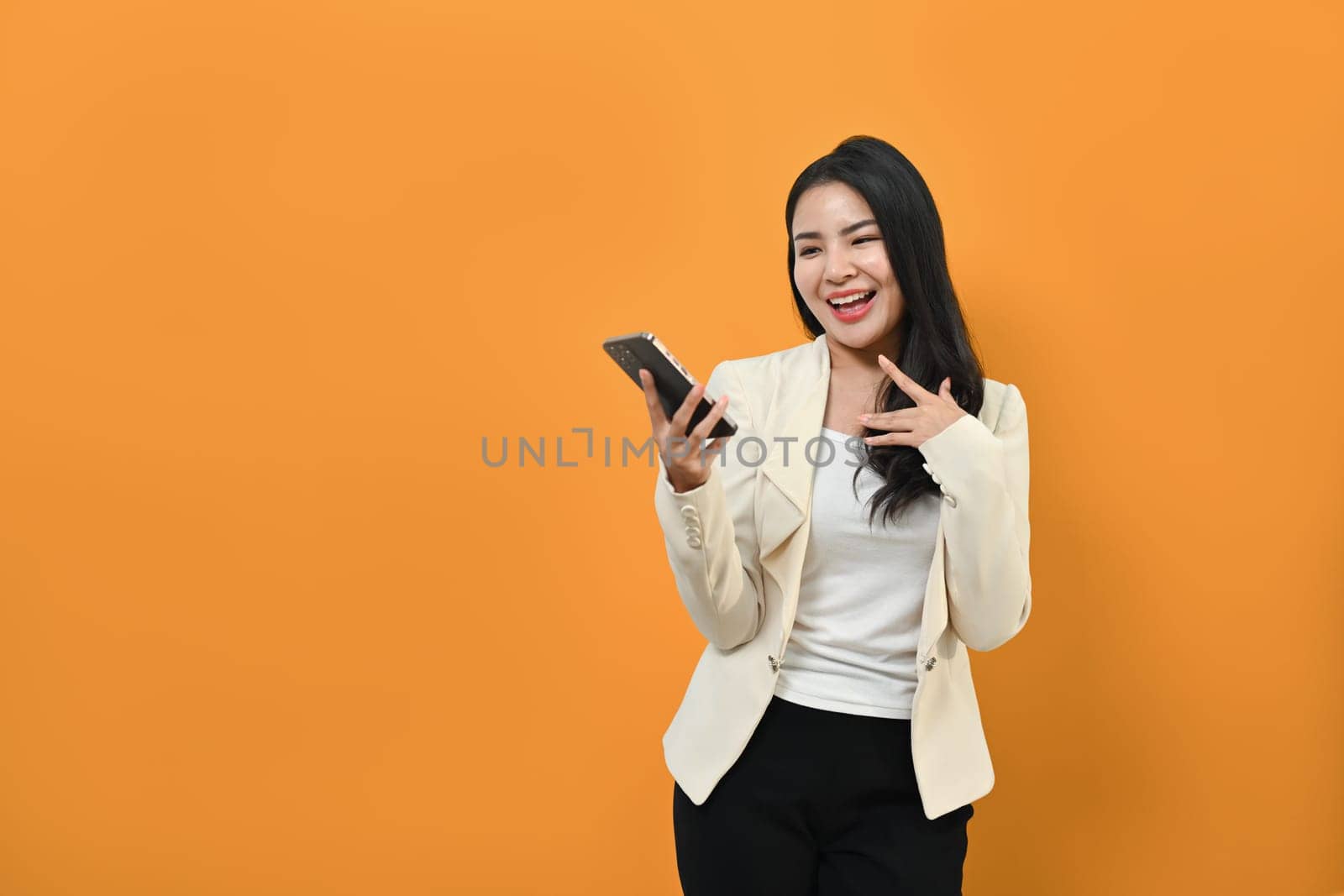 Cheerful asian woman in business suit holding smart phone in hands, standing on yellow background by prathanchorruangsak