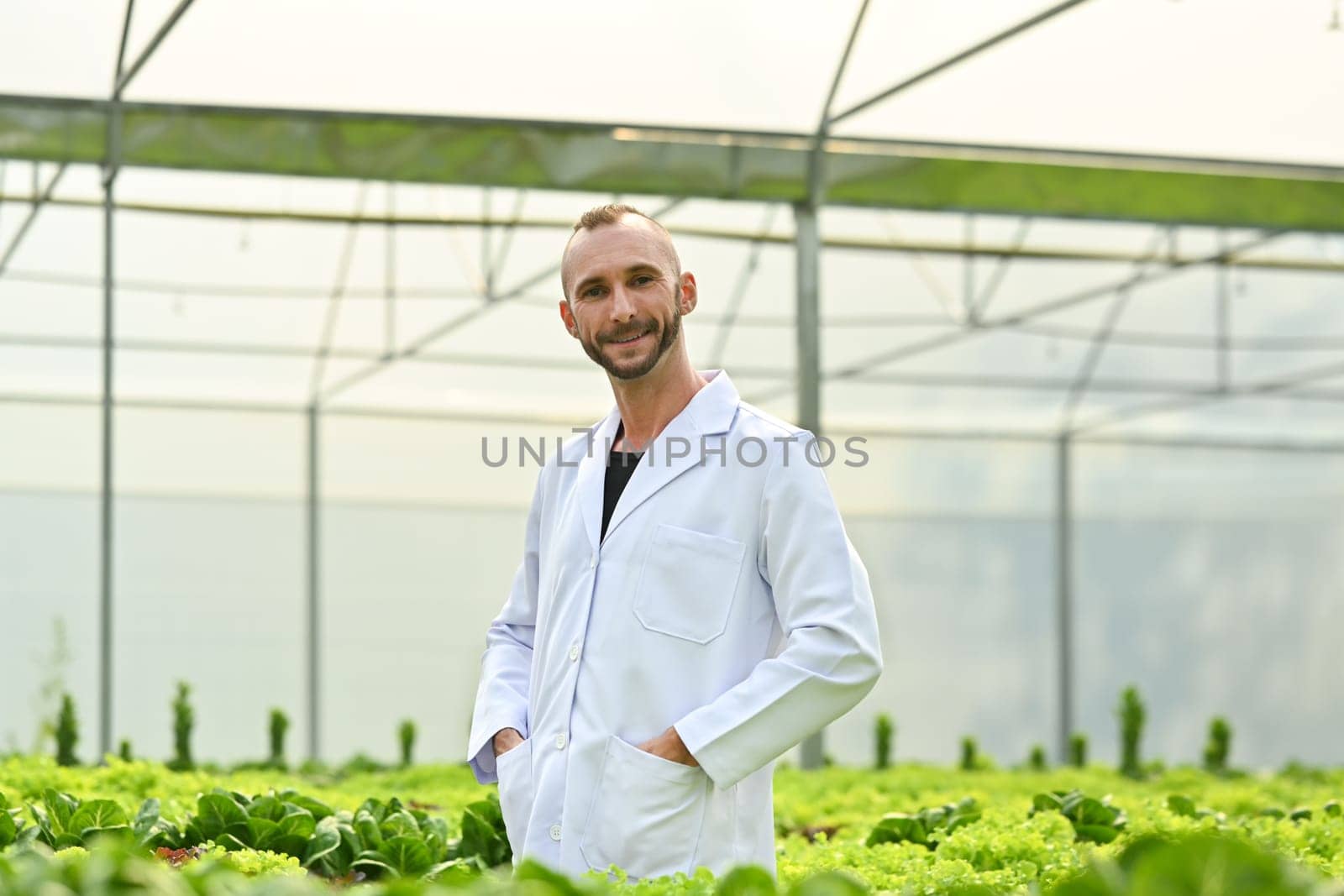 Positive caucasian male plant researcher, geneticist, biologist in white coat standing among vegetable in industrial greenhouse by prathanchorruangsak