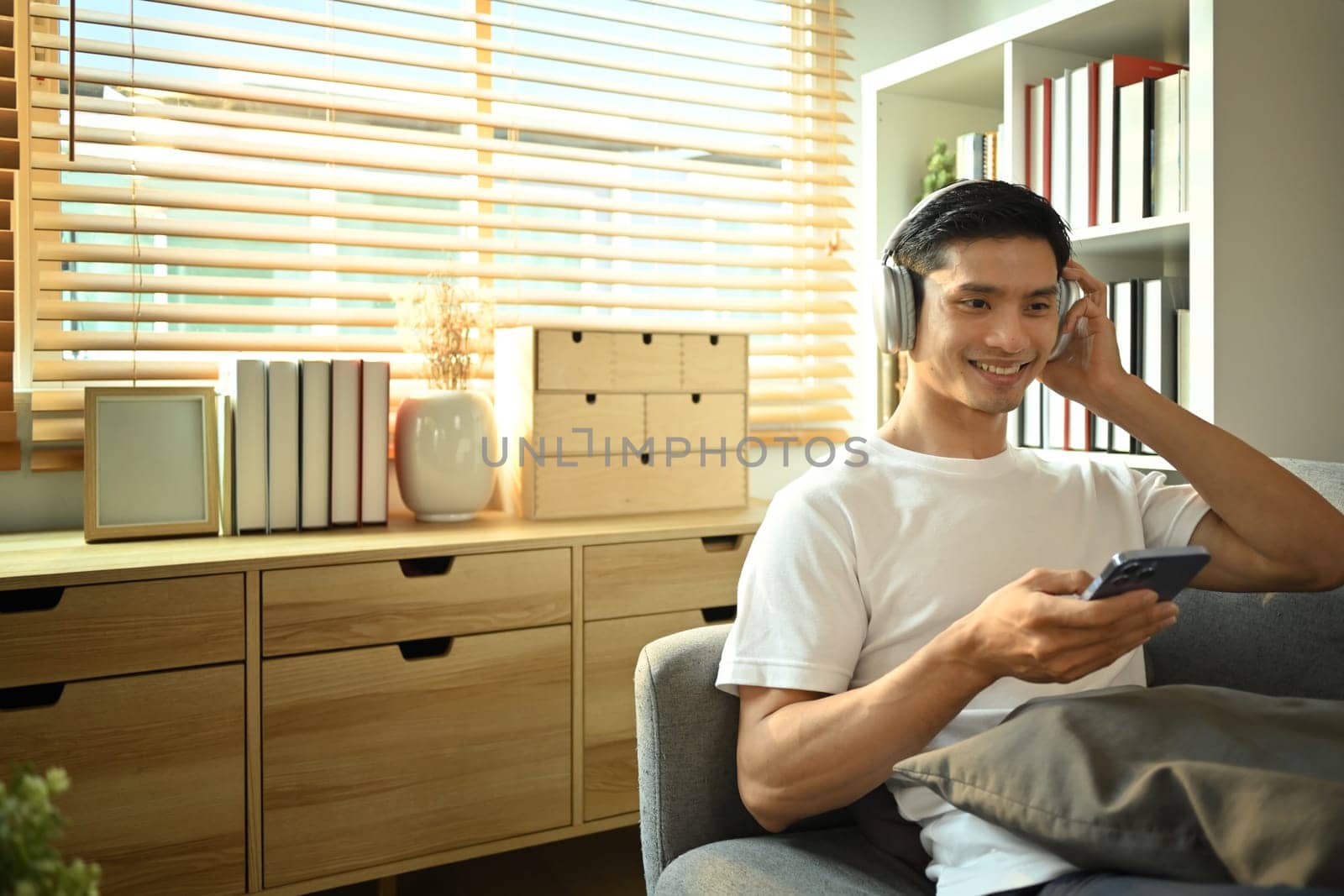 Happy adult asian man wearing wireless headphone and using phone on couch in cozy living room by prathanchorruangsak