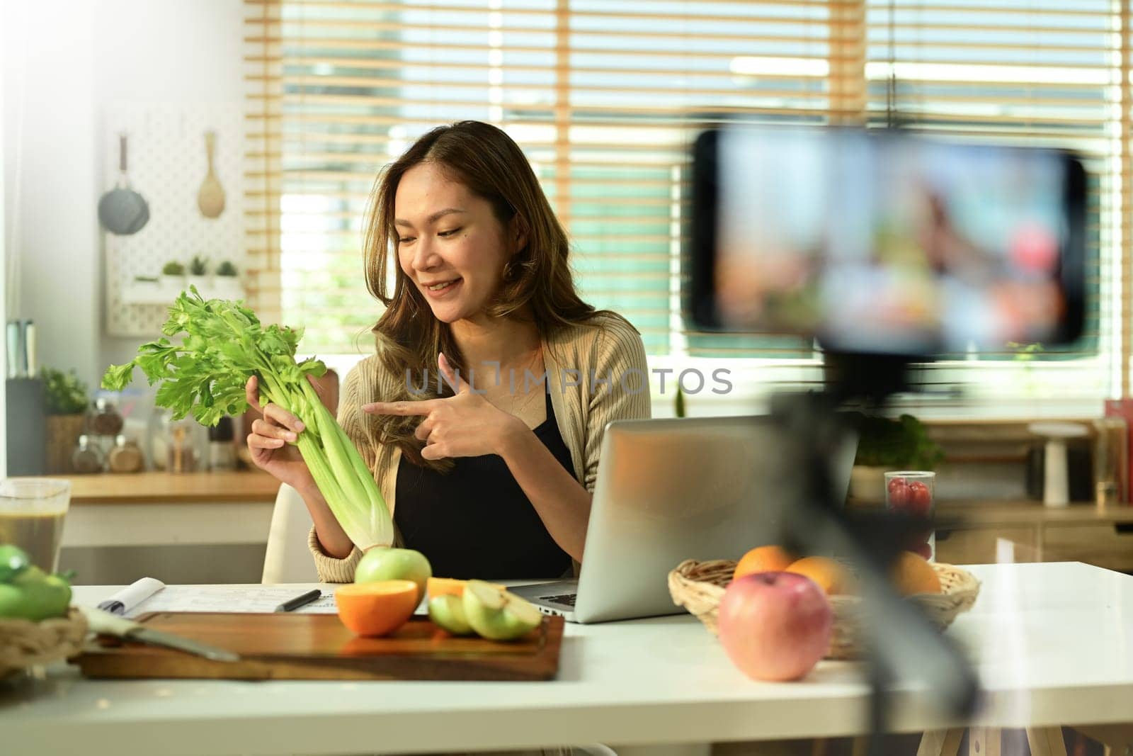 Female nutritionist blogger holding celery, providing with online consultations or recording video content on smartphone by prathanchorruangsak