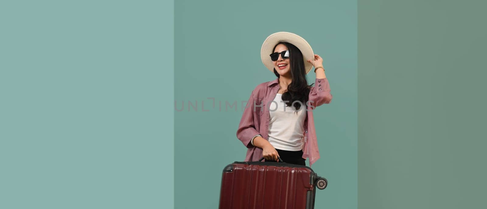 Happy Asian woman wearing summer hat and sunglasses with luggage standing isolated turquoise blue green background.