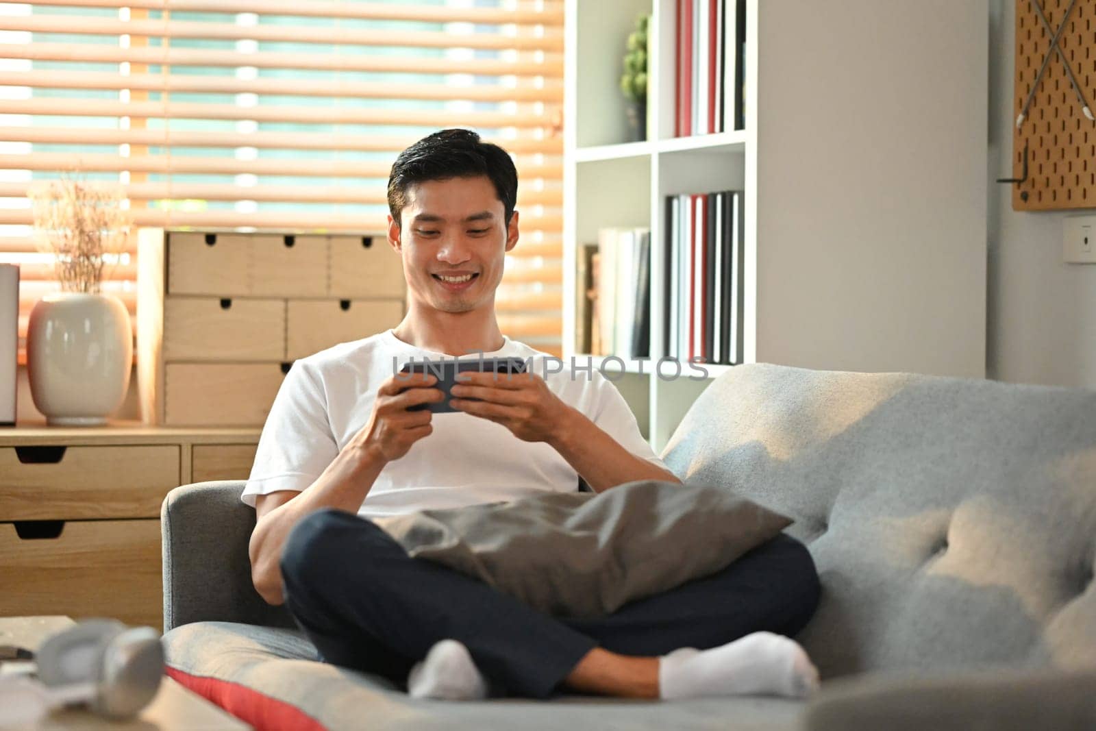 Joyful asian man in casual watching video or playing game on smart phone, sitting on couch at home.