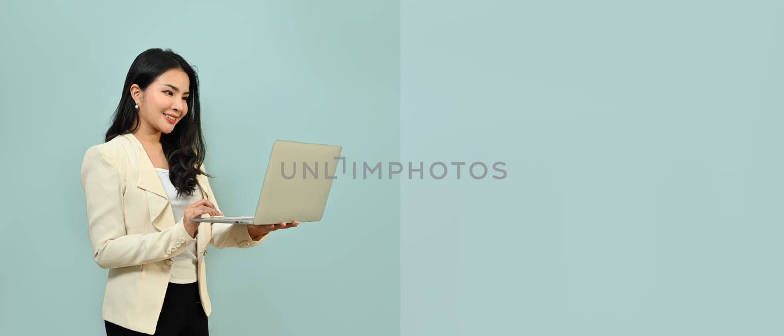 Beautiful asian woman in business clothes holding laptop computer standing isolated over blue background with copy space by prathanchorruangsak