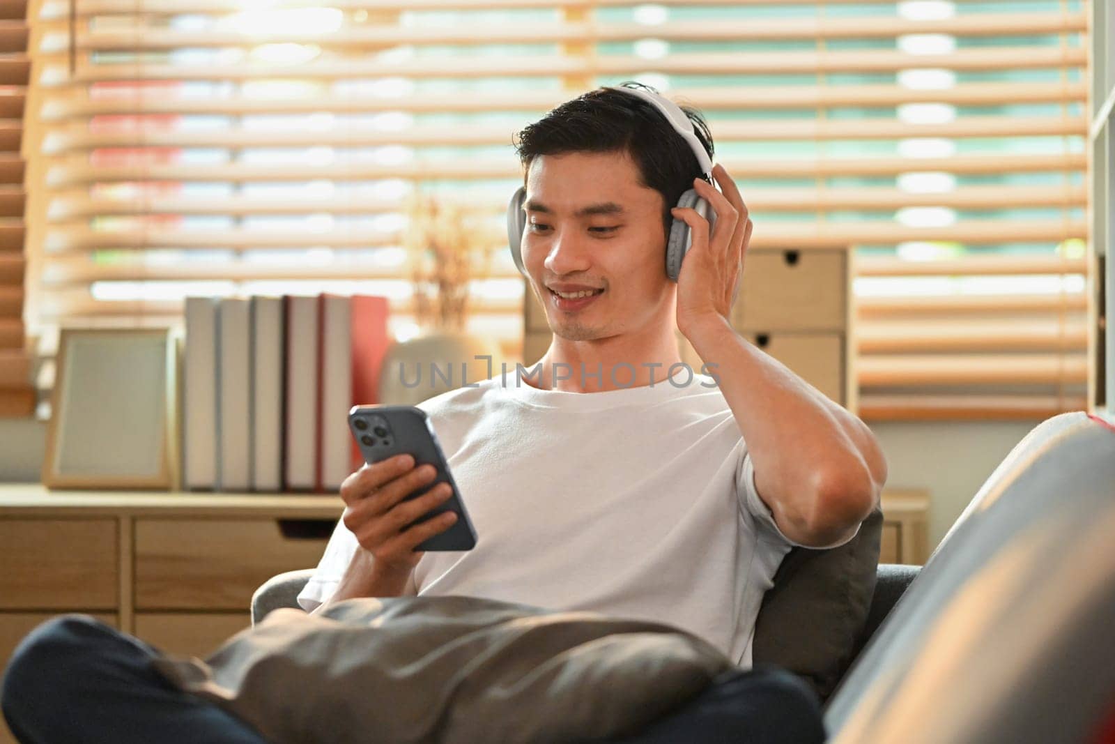 Handsome asian man in casual clothes listening to music in headphone and using smart phone on couch.