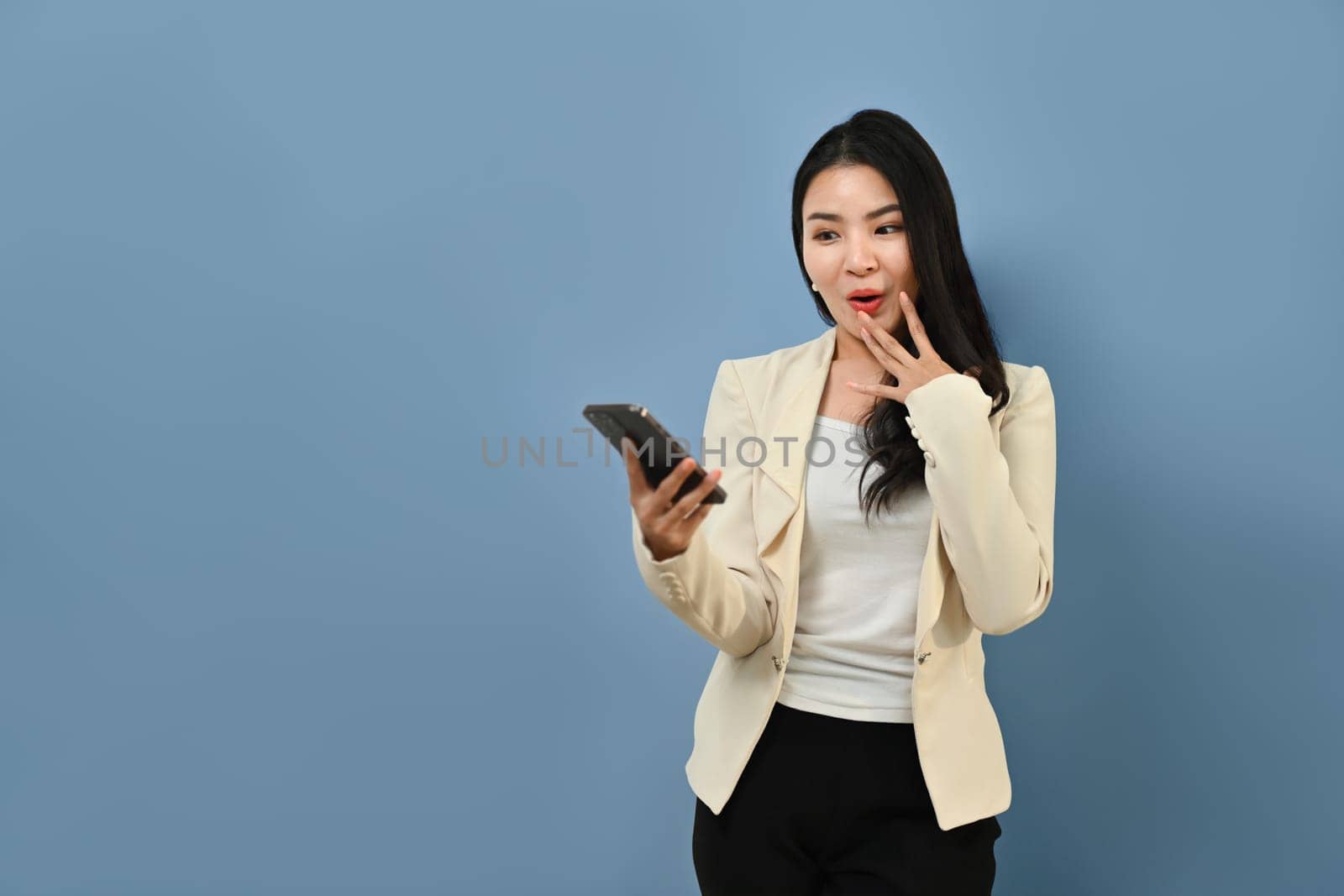 Surprised young Asian woman in stylish suit holding smart phone in hands, standing on blue background by prathanchorruangsak