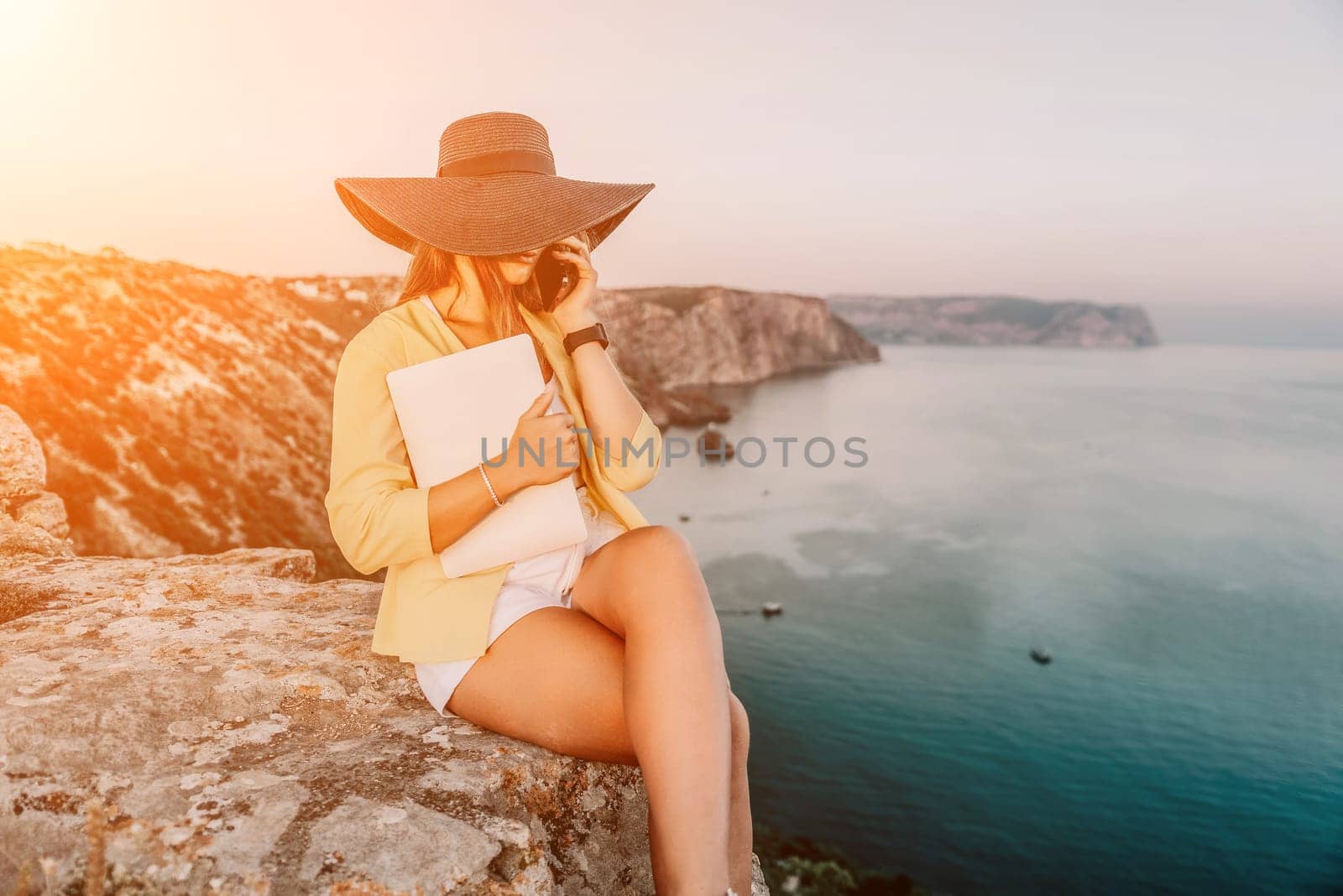 Successful business woman in yellow hat working on laptop by the sea. Pretty lady typing on computer at summer day outdoors. Freelance, travel and holidays concept.