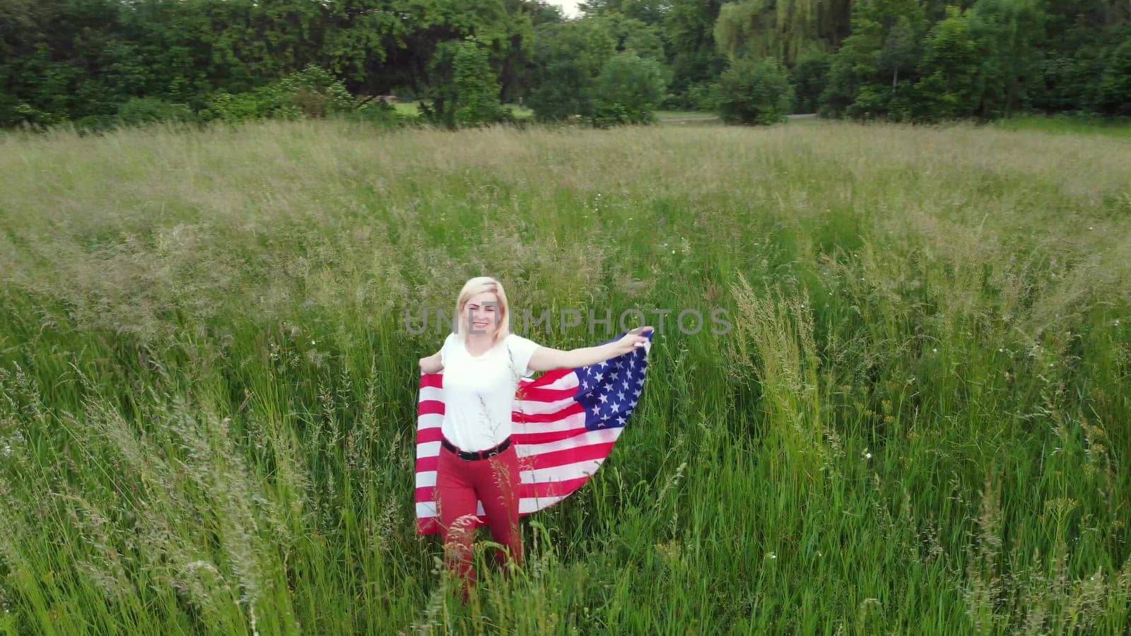 full length blond Woman holding American Flag in trees shadow of sunlight Back view cute blondy girl run on fresh green grass texture.