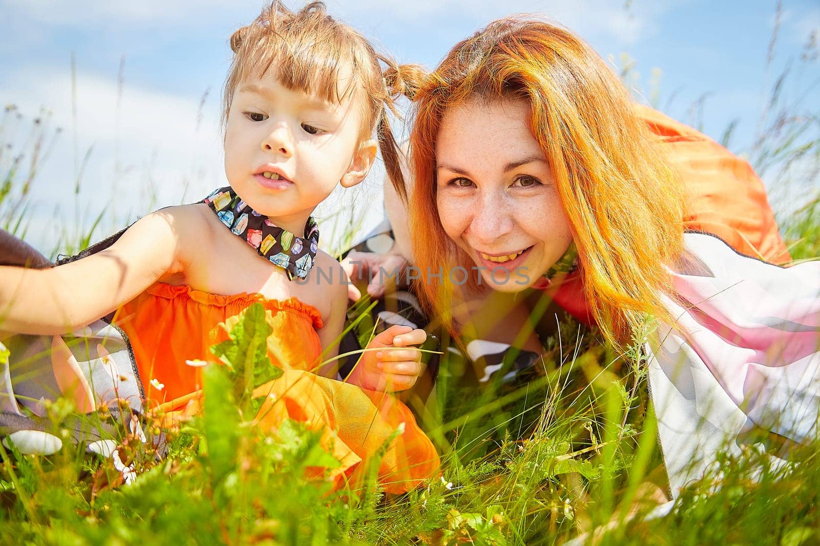 Happy female family with mother and daughter on green and yellow meadow full of grass and flower. Woman with red hair and blonde girl having fun, joy and hug in sunny summer day. Concept family love