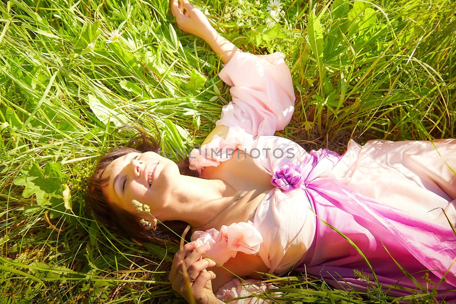 Beautiful girl in lush pink ball gown in green field during blooming of flowers and blue sky on background. Model posing on nature landscape as princess from fary tale