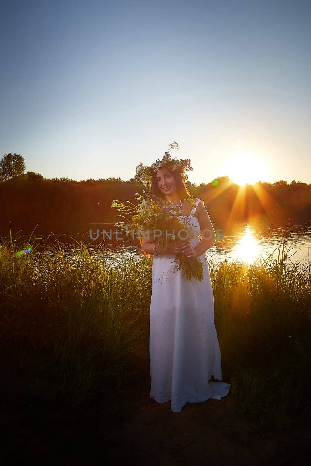 Adult mature brunette woman in a white dress, sundress and a wreath of flowers in summer by the water of river or lake in evening at sunset. Celebration of the Slavic pagan holiday of Ivan Kupala