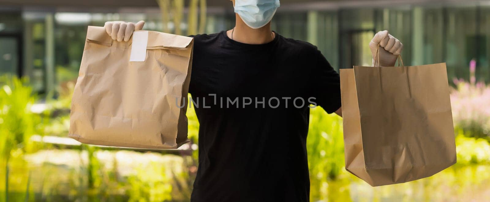 Delivery guy with protective mask and gloves holding bag with groceries in front of a building