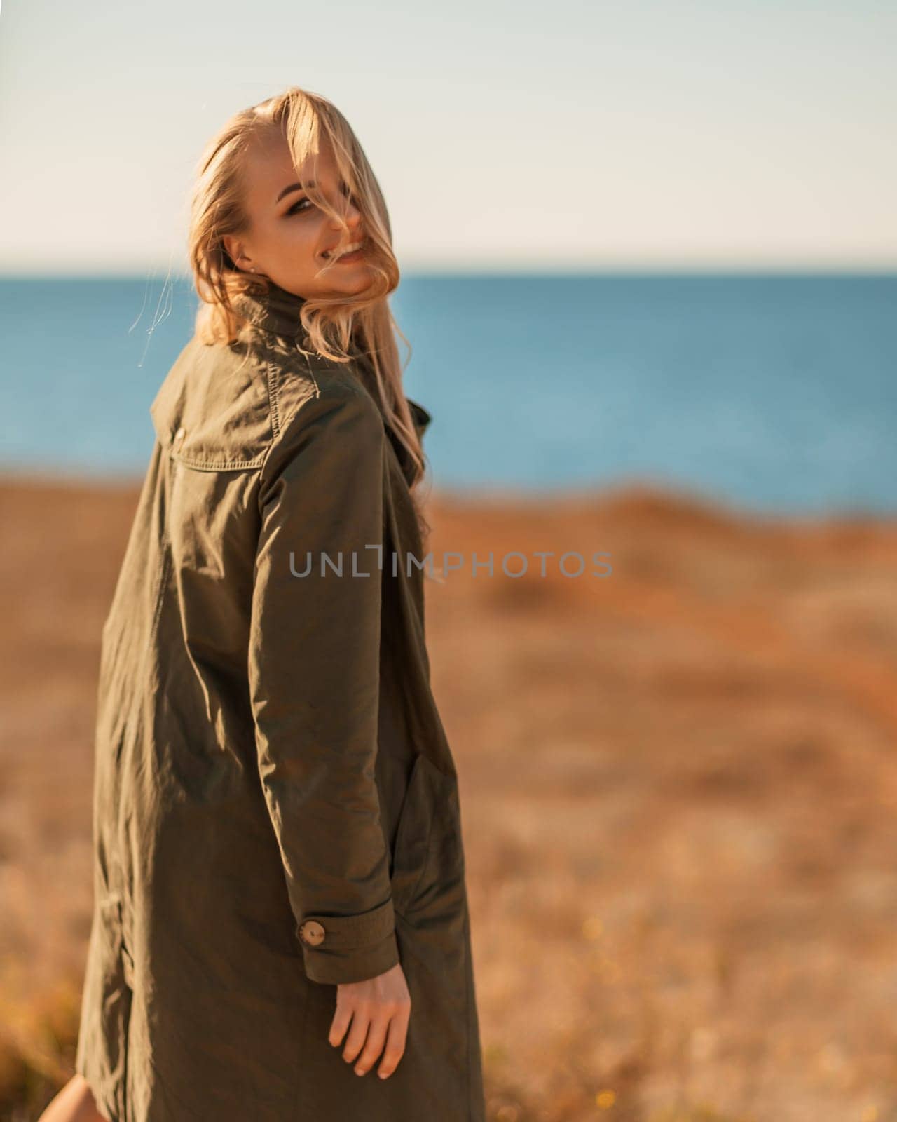 Portrait blonde sea cape. A calm young blonde in an unbuttoned khaki raincoat stands on the seashore, under the raincoat there is a black skirt and top by Matiunina