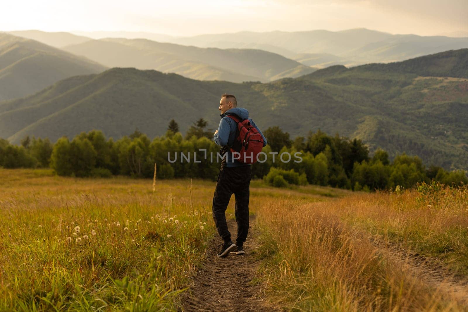 Guy tourist with a backpack stands in the mountains on a meadow with a backpack on his back.