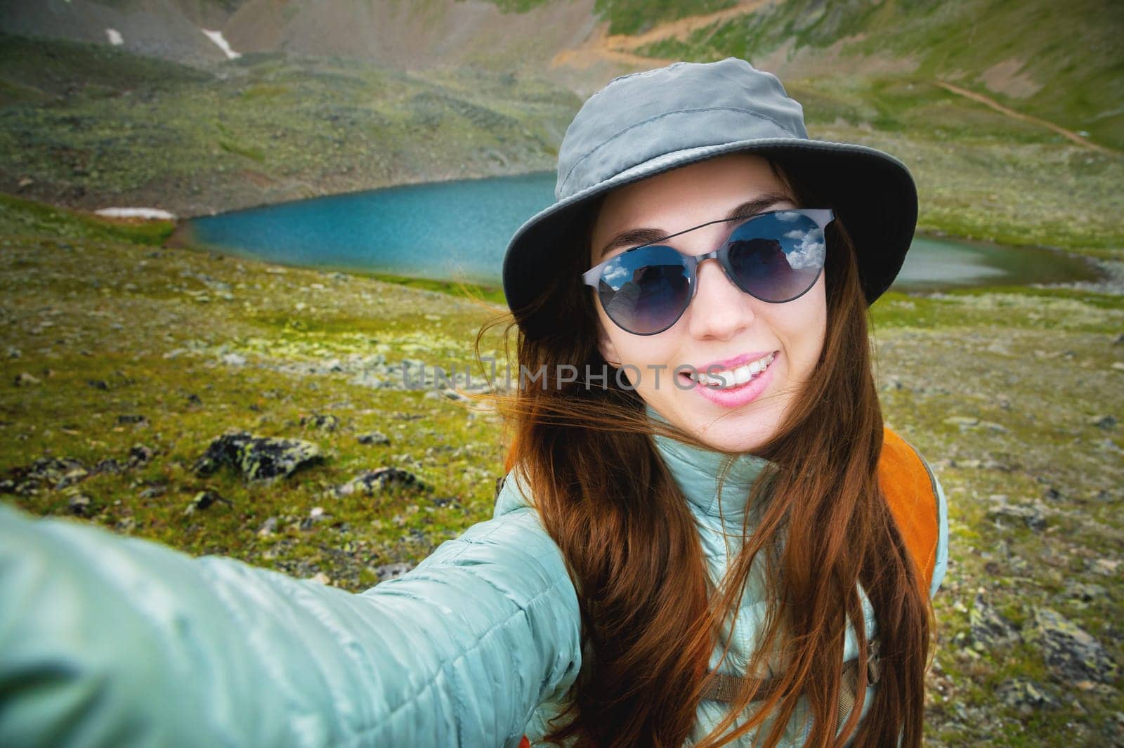 Portrait of a young happy smiling woman with long brunette hair, wearing a jacket and sunglasses, taking a selfie over the top of a mountain lake. Adventure, travel, vacation concept.