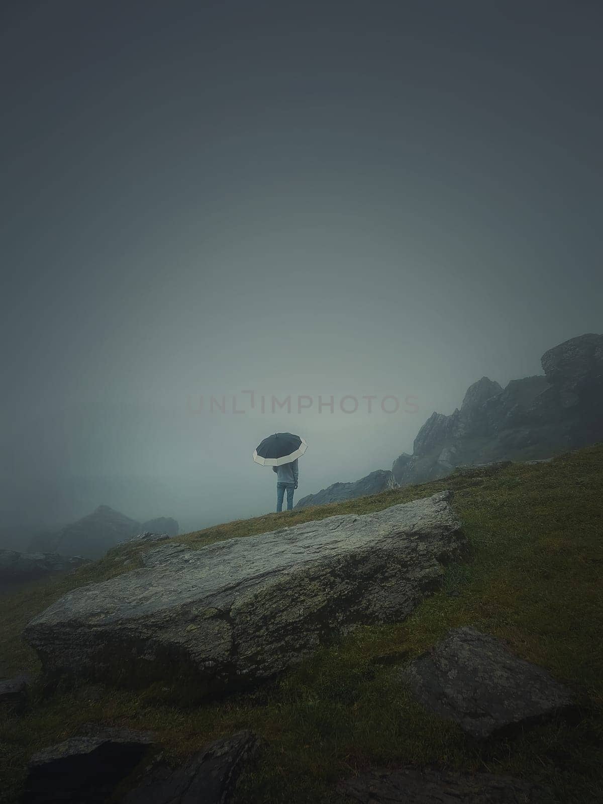 Rear view of a lonely man with umbrella stands on a rocky hill covered by haze. Moody and emotional scene with a lone stranger silhouette under the rain by psychoshadow