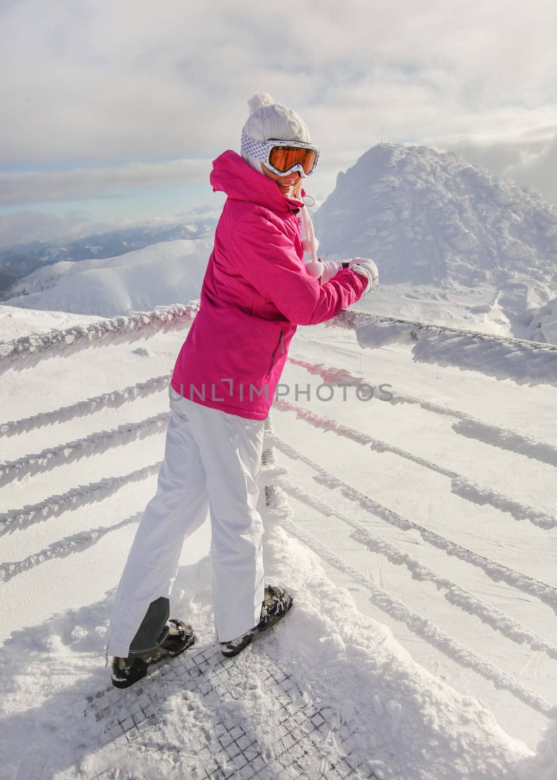 Young woman in pink ski jacket, pants boots, hat and gloves, leaning on snow covered rail, looking back, smiling.