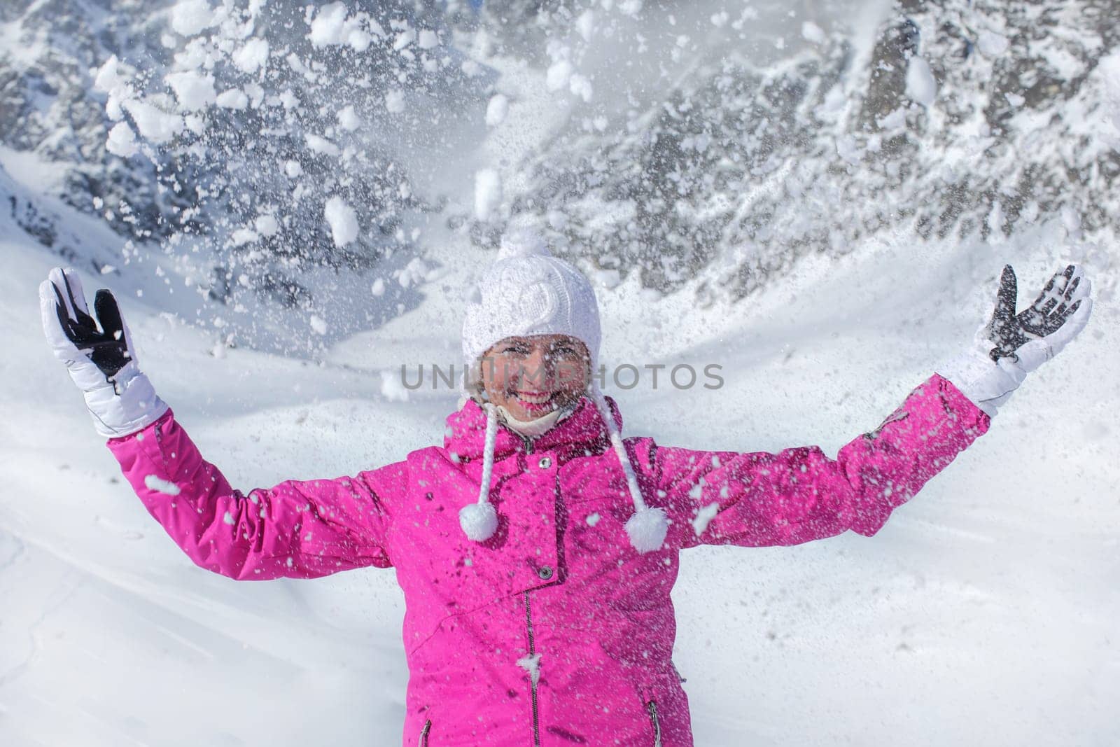 Young woman in pink ski jacket, gloves and winter hat, smiling, throwing snow in the air by Ivanko