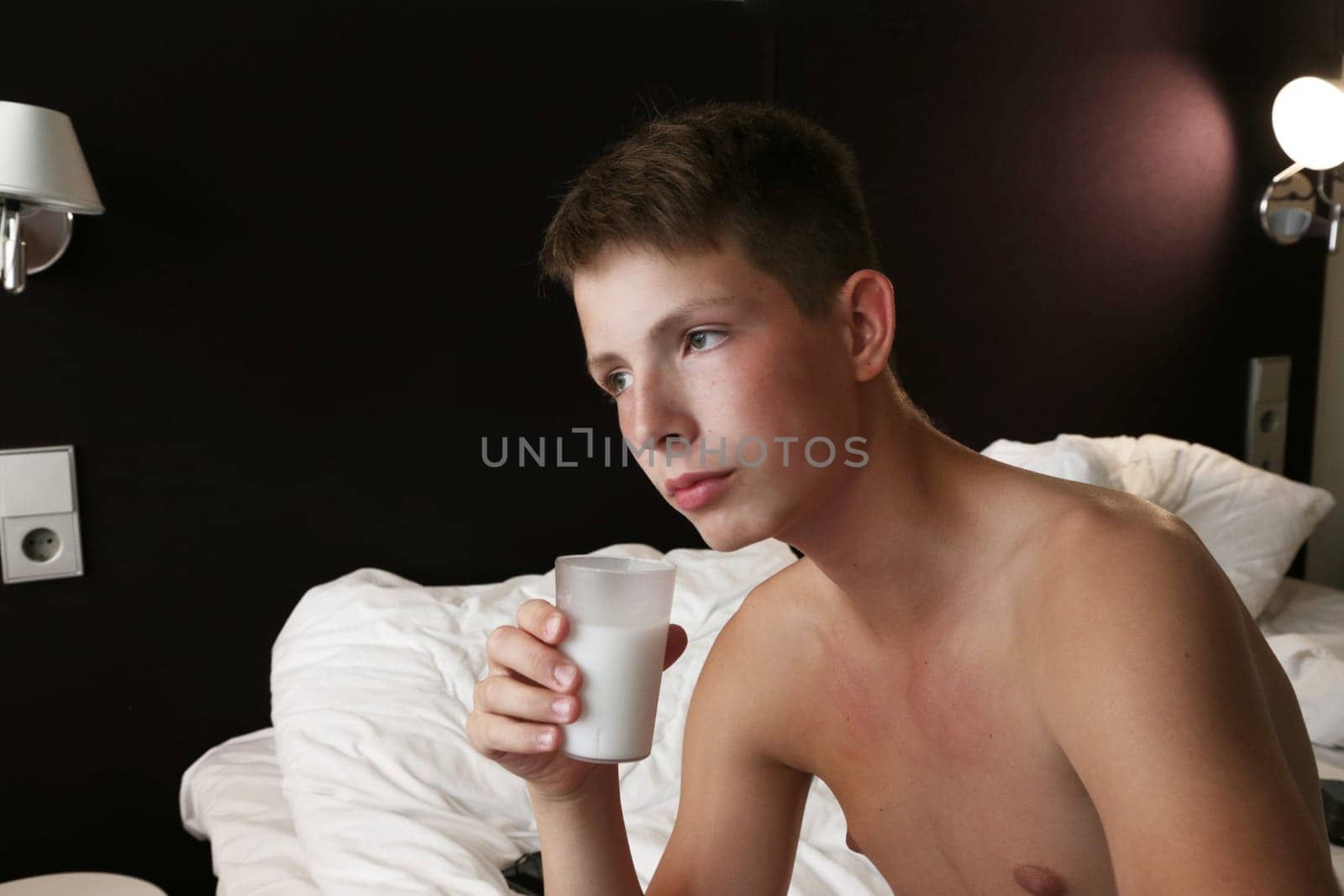 A cute boy sits on the bed and holds a glass in his hand. A teenage boy sits on a white bed in a hotel room and holds a glass of milk in his hand.