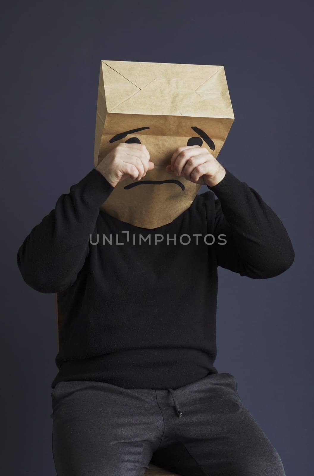 A sad man in a black turtleneck with a bag on his head, with a drawn crying emoticon, cries and wipes away a tear. Emotions and gestures. Vertical frame.