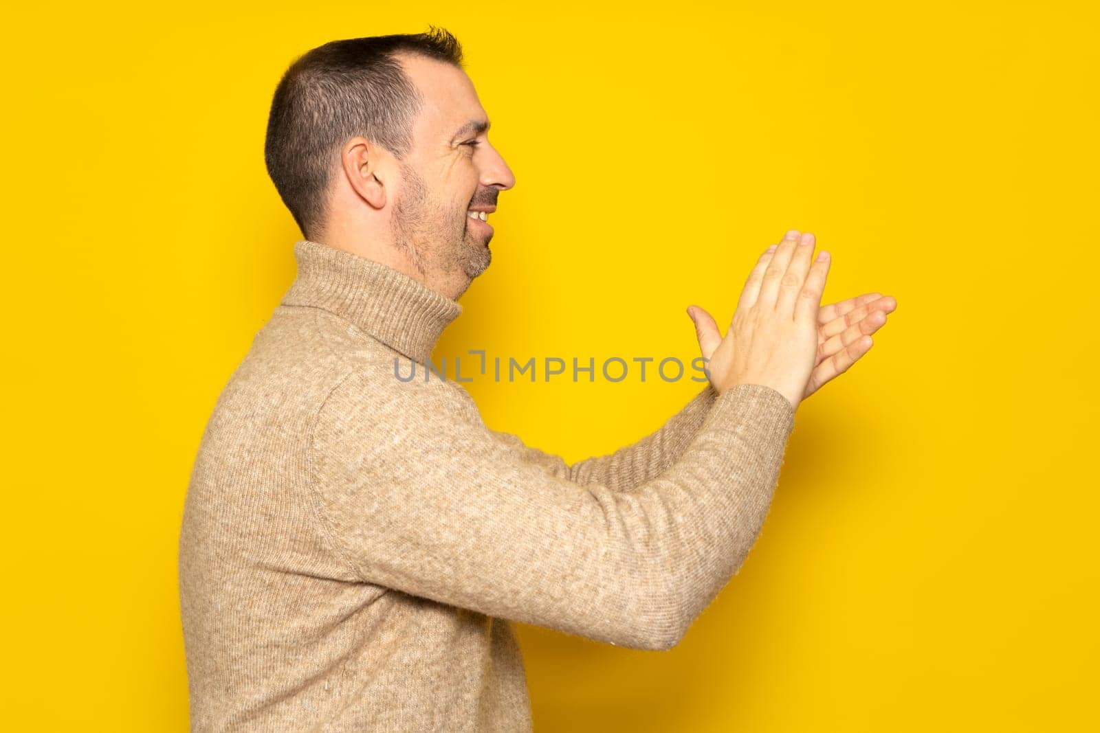 Bearded hispanic man in profile wearing brown turtleneck feeling happy, smiling and clapping hands with emphasis thankful for the great show he just witnessed. Isolated on yellow studio background