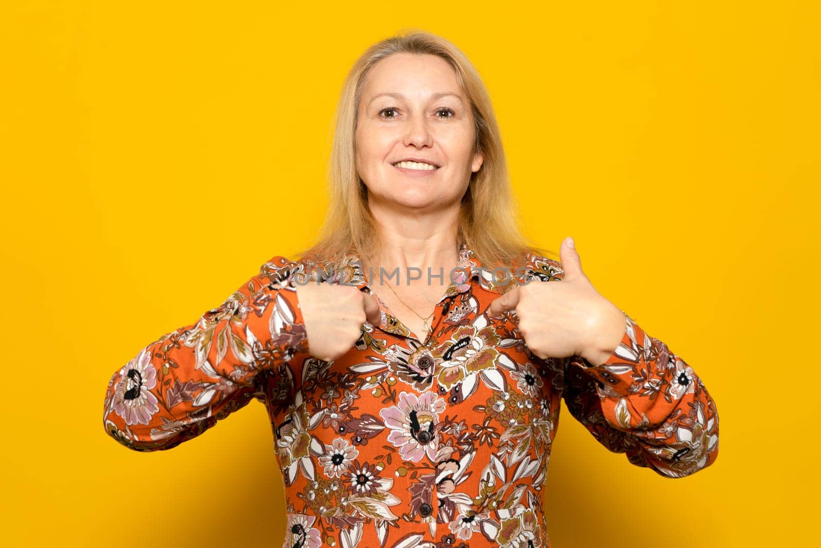 Caucasian blonde woman in her 40s in a patterned dress pointing to herself proud of being the way she is, strong and independent woman isolated over yellow background