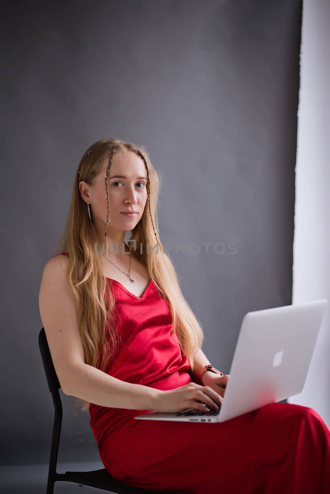A business woman blogger in a red dress is working, typing on a laptop apple and sitting on a chair. portrait blonde assistant of hands with computer MacBook . Vertical