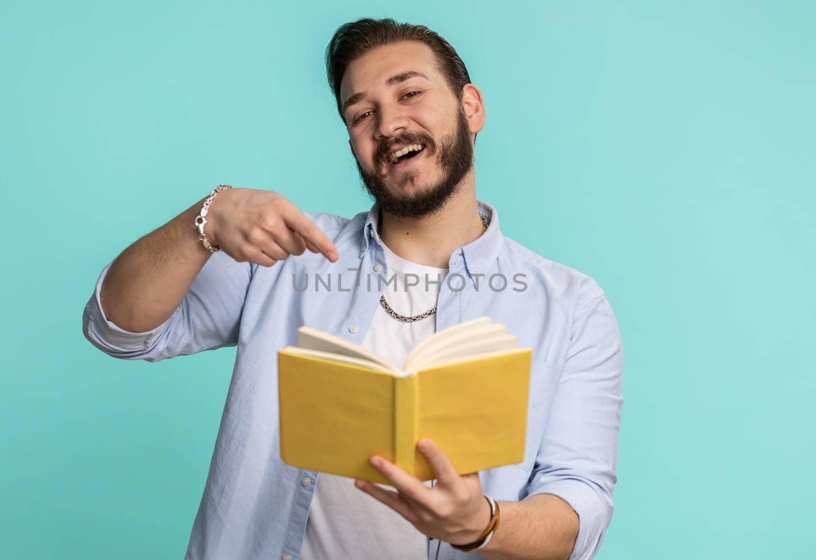 Happy lebanese man reading funny interesting fairytale story book, leisure hobby, knowledge wisdom, education, learning, study, wow. Handsome arabian guy isolated alone on blue studio wall background