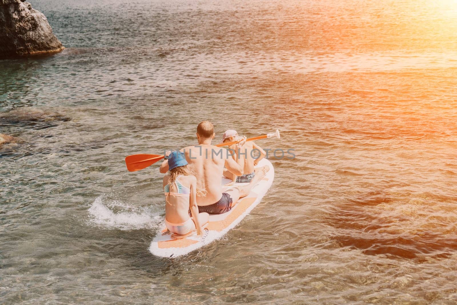 Father and his adorable little son and daughter sitting on stand up board having fun during summer beach vacation