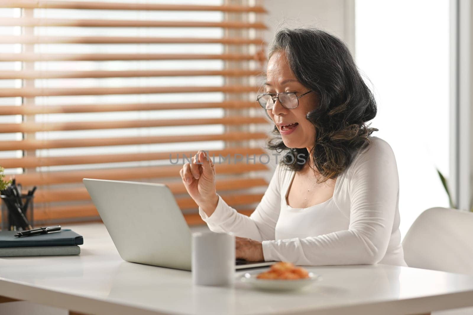 Attractive senior woman in glasses watching online webinar on laptop during remote working from home.