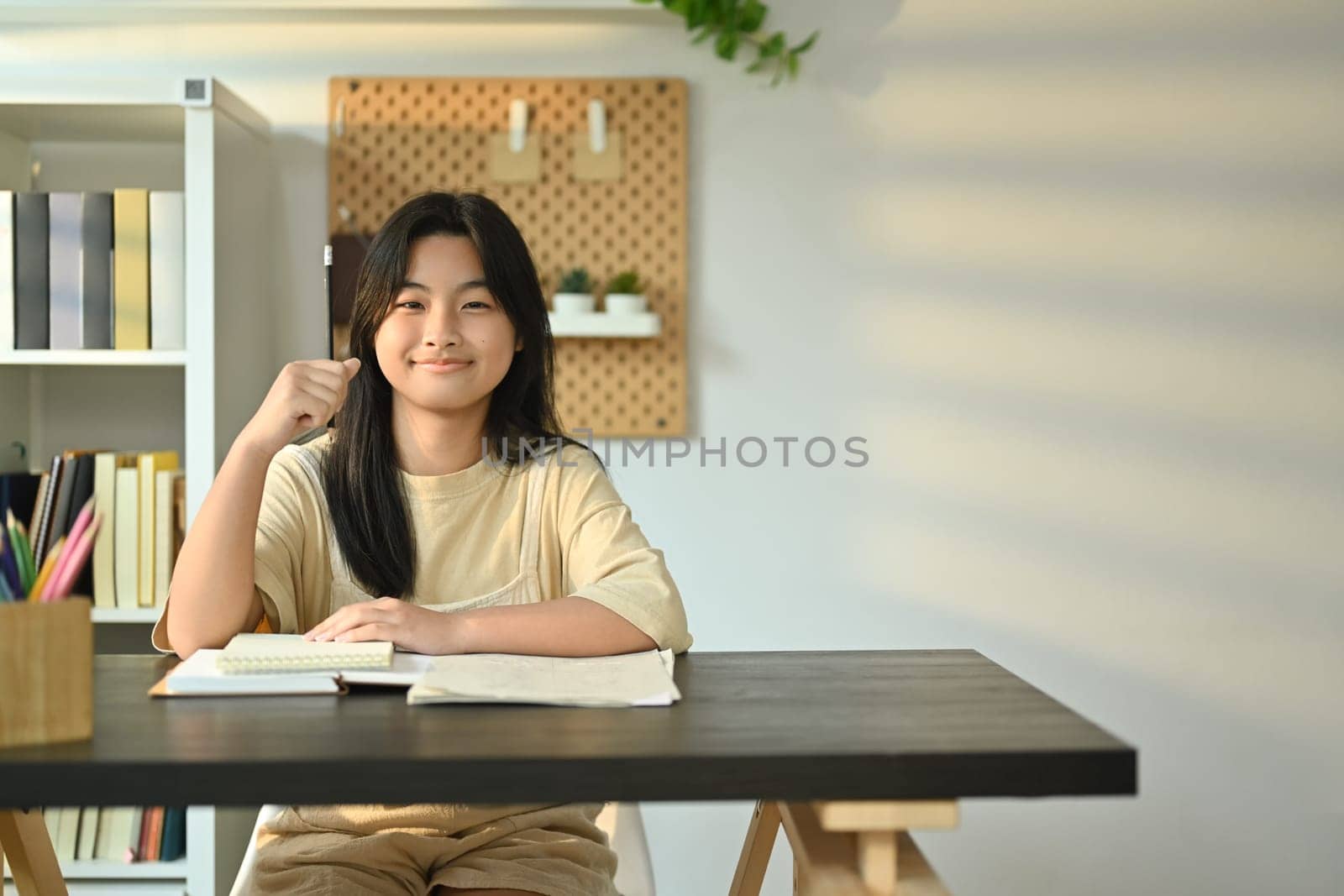 Cute teenager girl with a lovely engaging smile sitting at a desk in living room. Homeschooling, learning, self education by prathanchorruangsak