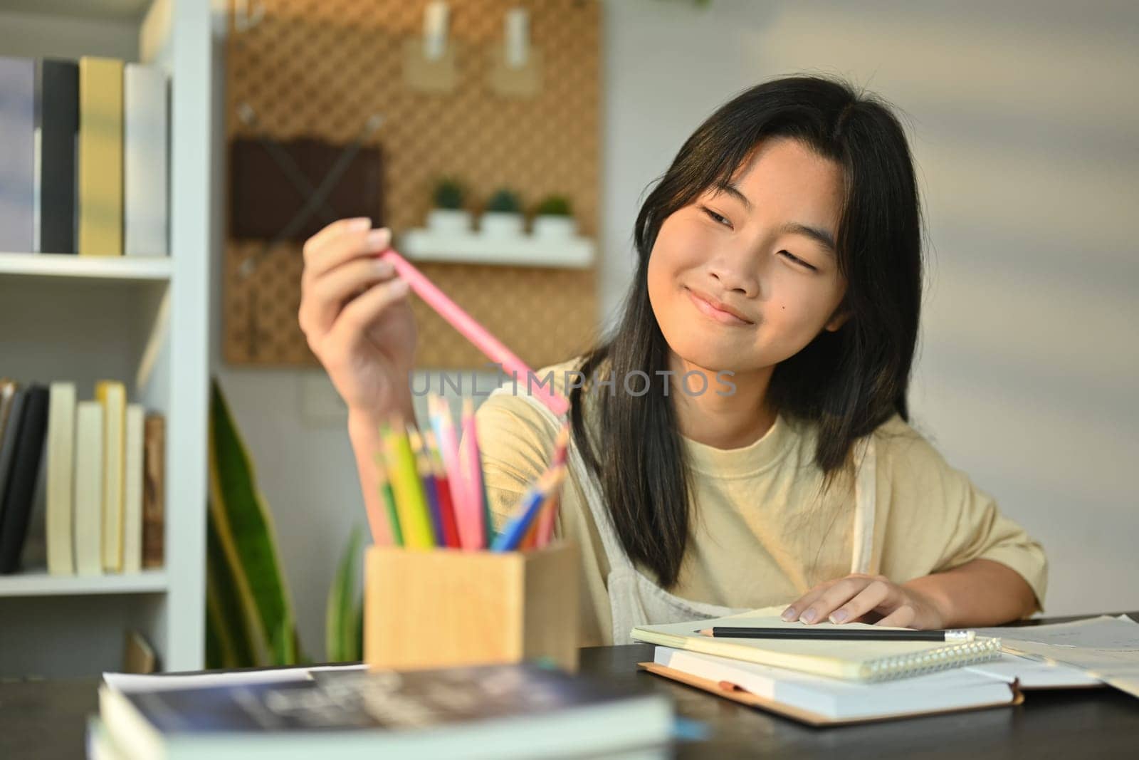 Cheerful teenaged girl doing homework, writing in notebook while sitting at table in living room by prathanchorruangsak