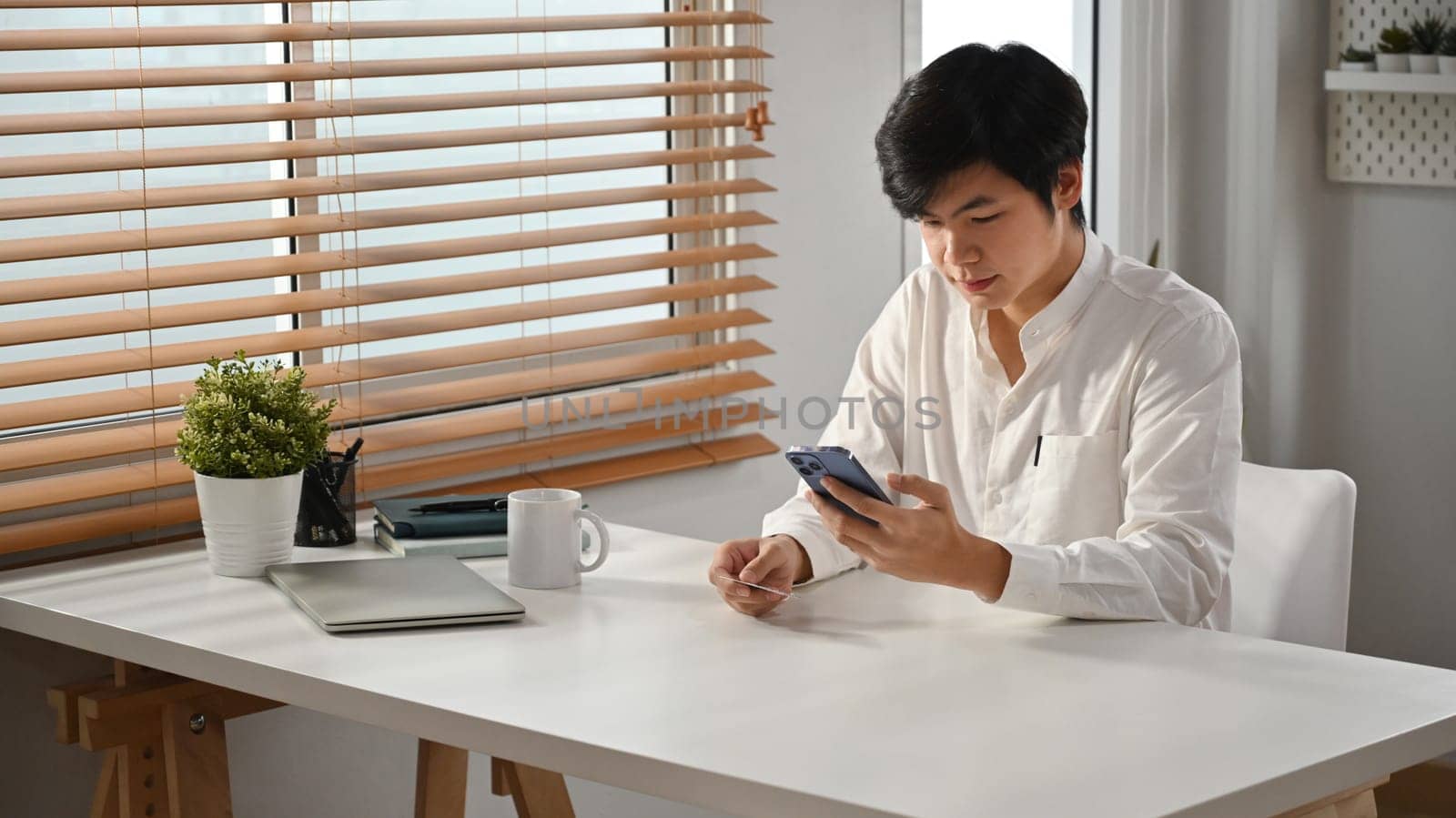 Millennial asian man sitting in modern home office and using smartphone. People, Technology and Communication.