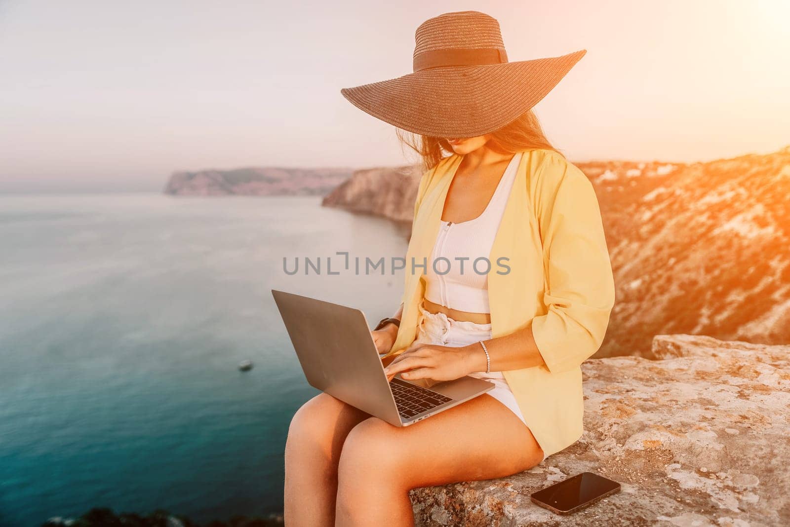 Successful business woman in yellow hat working on laptop by the sea. Pretty lady typing on computer at summer day outdoors. Freelance, travel and holidays concept.