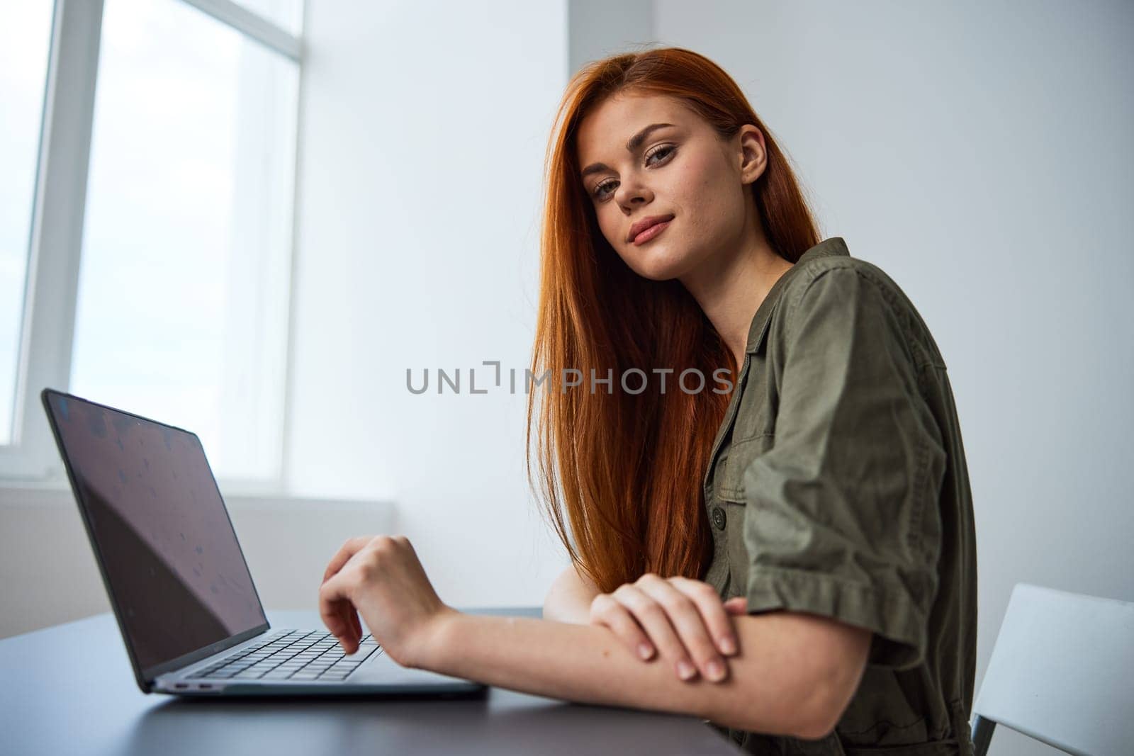 portrait of a cute, smiling red-haired woman sitting at a laptop with her hands folded on the table and looking at the camera by Vichizh