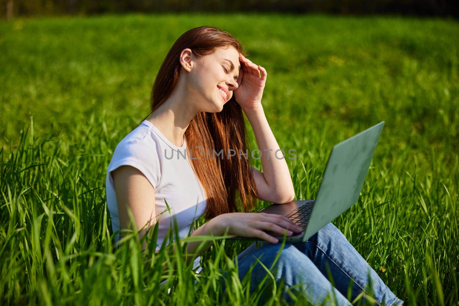 a woman works at a laptop sitting in a field covering her face from the sun with her hand by Vichizh