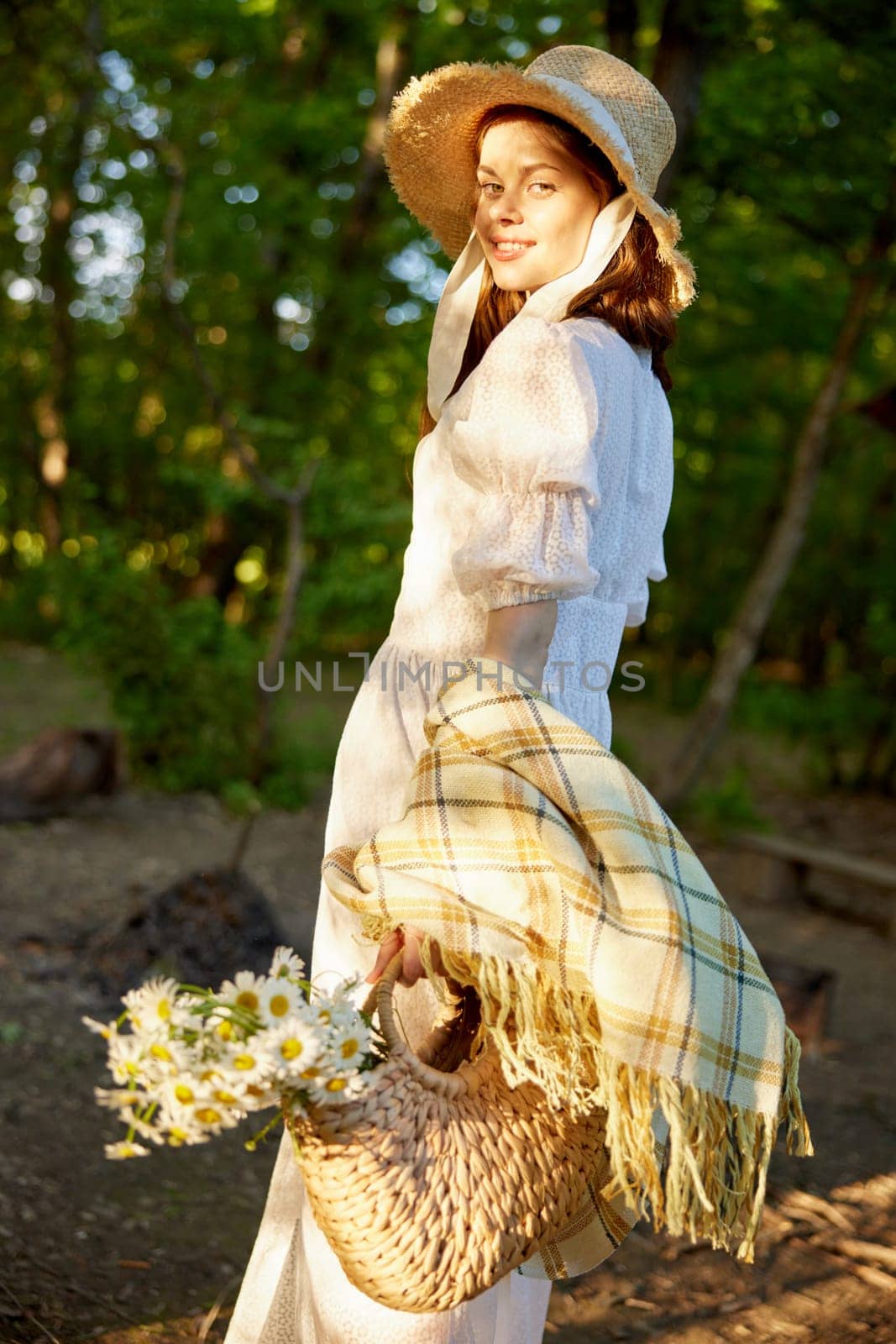 a woman in a wicker hat walks through the forest and looks towards the sunset holding a plaid and a bag in her hands. High quality photo