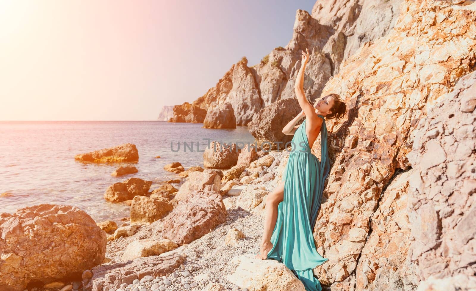 Side view a Young beautiful sensual woman in a red long dress posing on a rock high above the sea during sunrise. Girl on the nature on blue sky background. Fashion photo.