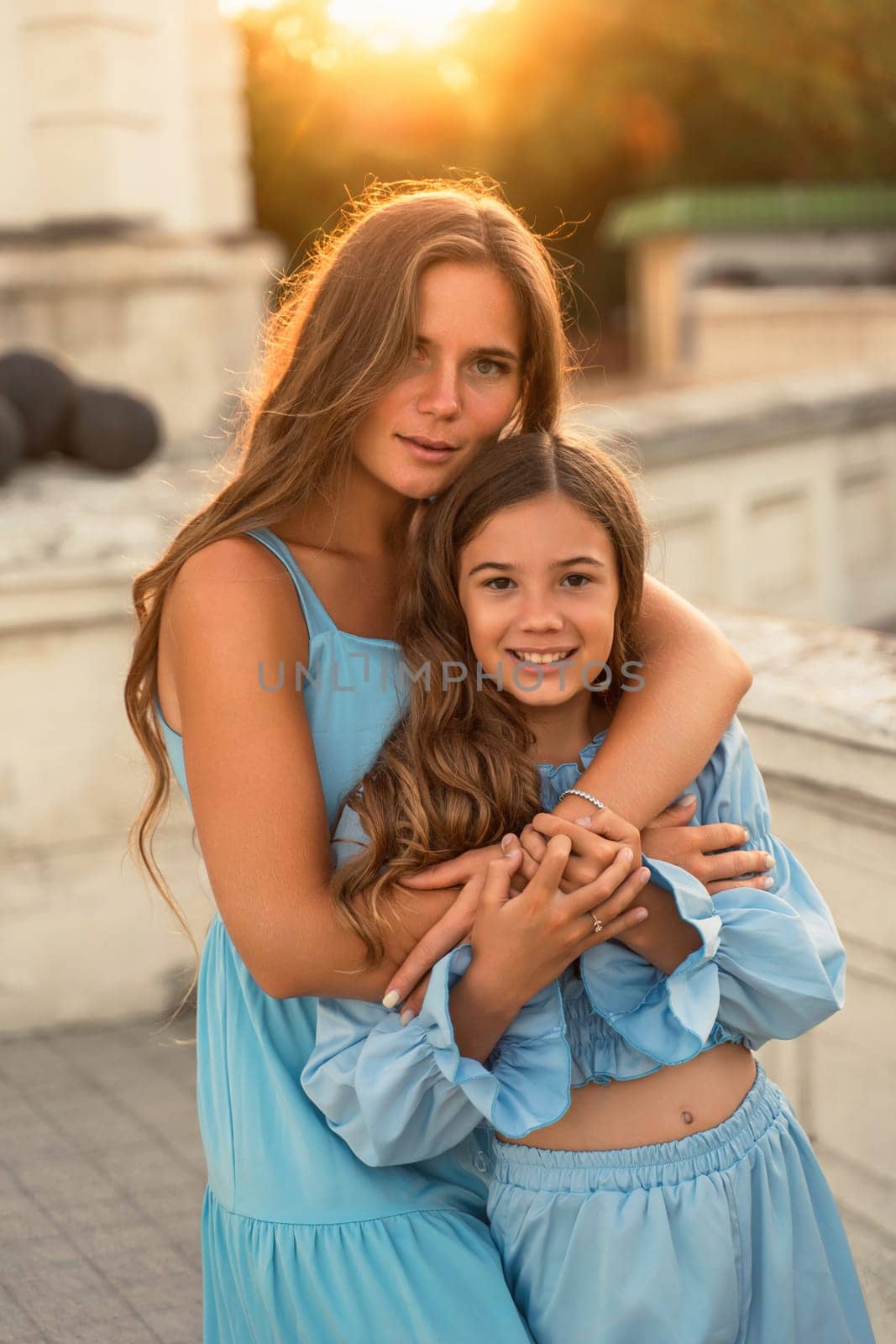 Portrait of mother and daughter in blue dresses with flowing long hair against the backdrop of sunset. The woman hugs and presses the girl to her. They are looking at the camera