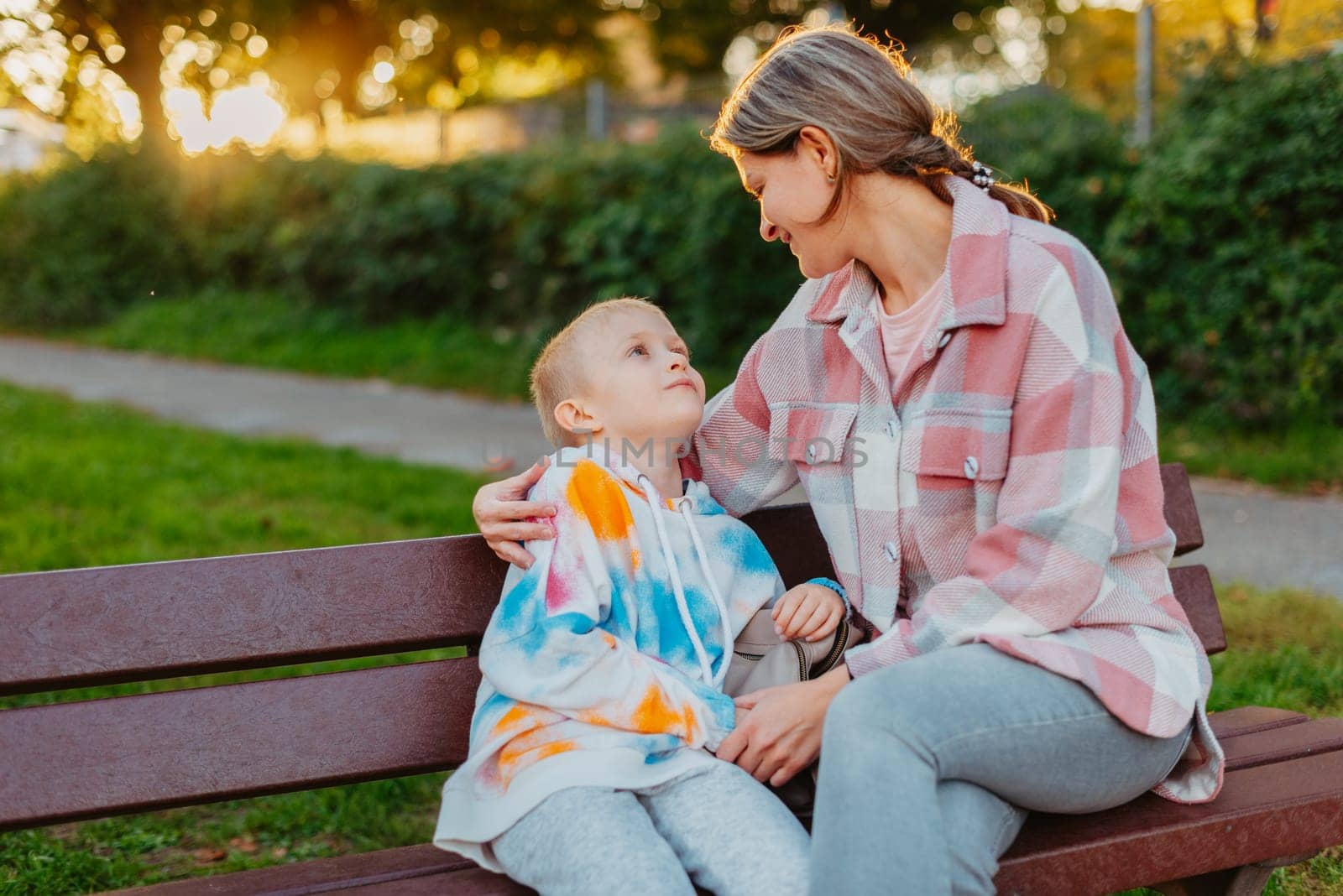 mother and son sit on a park bench in the rays of the setting sun. the concept of a family. Mother's Day. beautiful girl (mother) with a boy (son) in the park in the park are sitting on a bench at sunset by Andrii_Ko