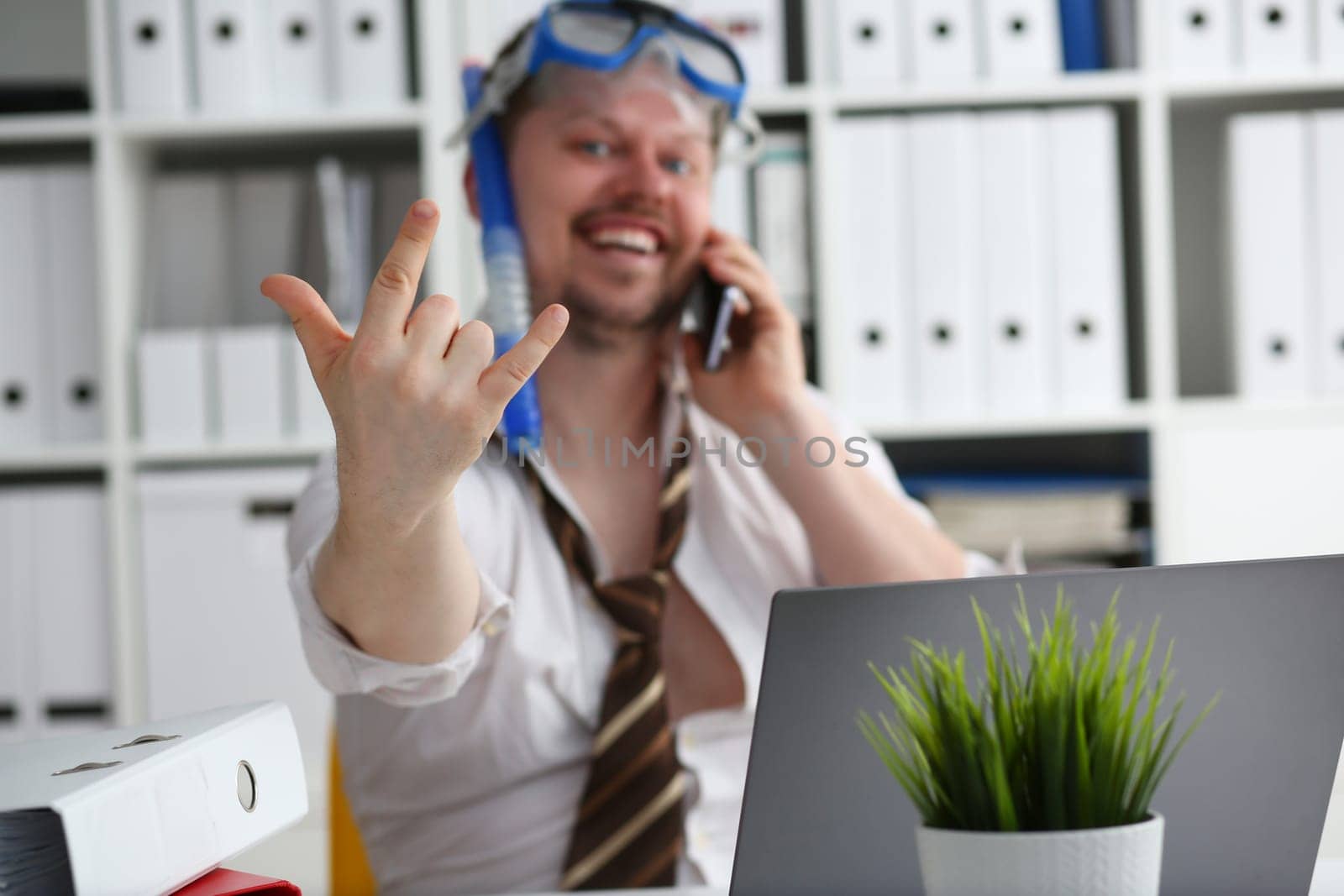 Emotionally cheerful male manager in travel company in swimming goggles is talking on mobile phone at workplace. Sale hot tourist tours travel and vacation concept