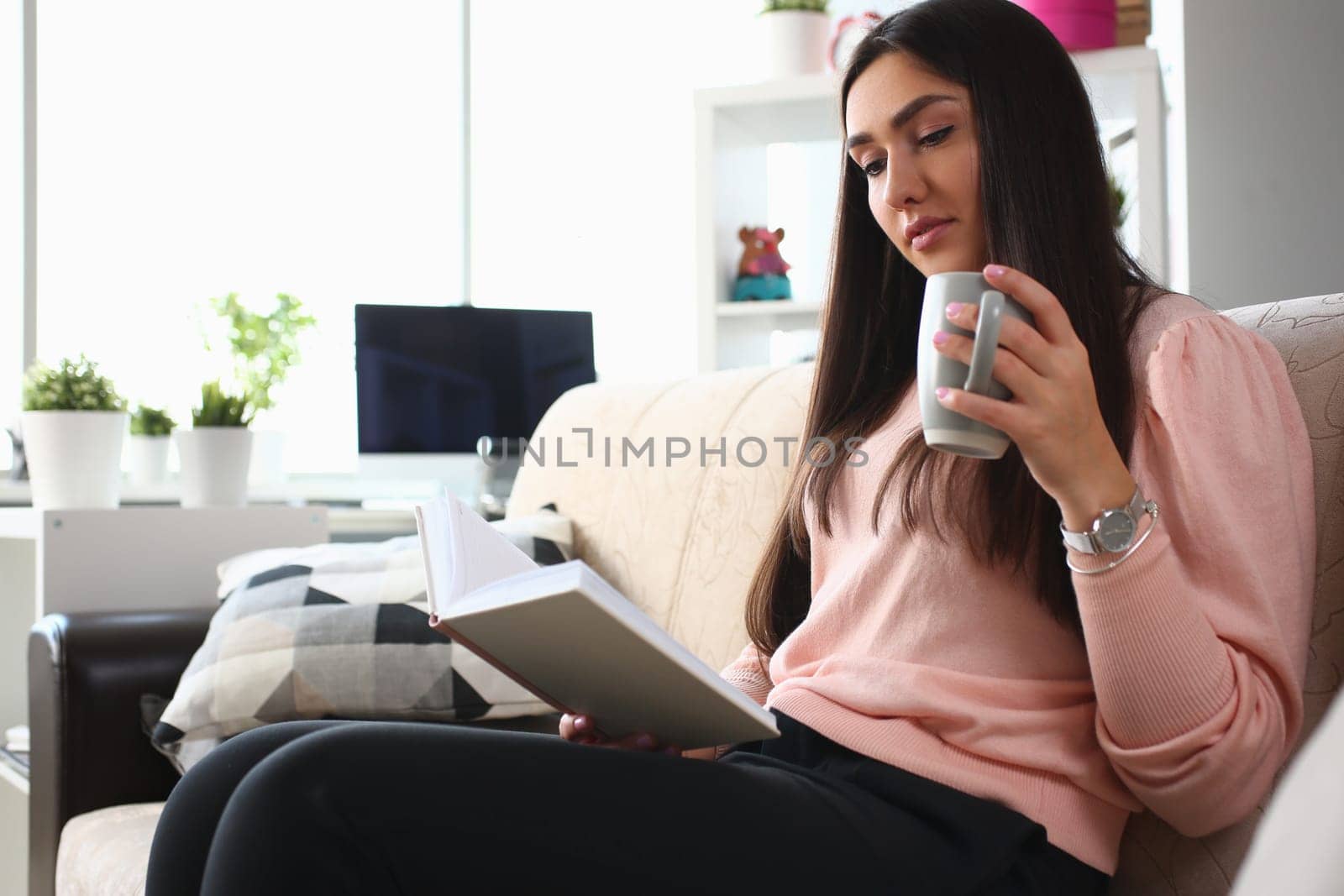 Woman is reading book and drinking from cup sitting on couch. Home leisure and reading books concept