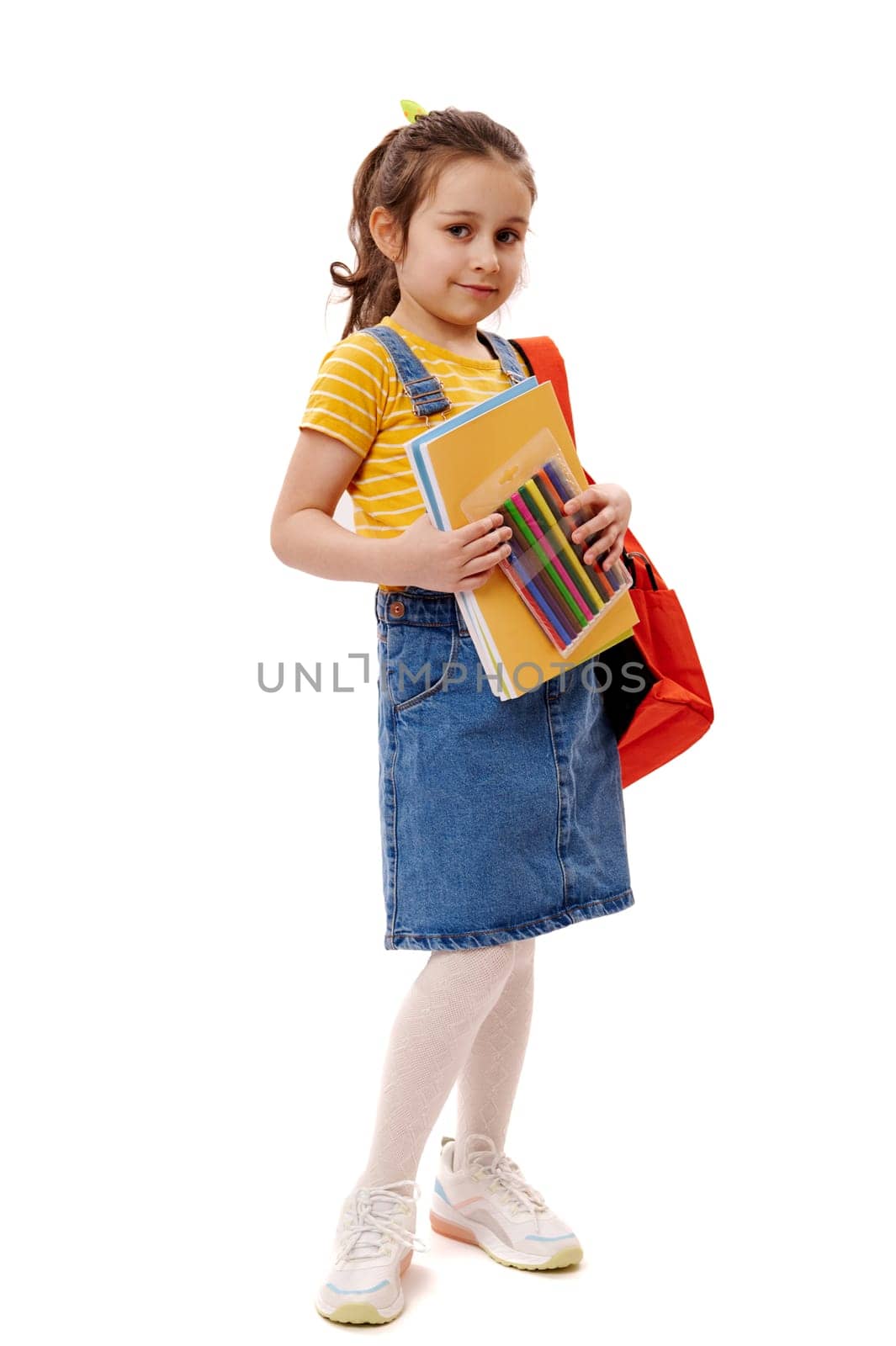 Full length portrait of 5-6 years child, smart schoolgirl in yellow t-shirt and blue denim sundress, carrying backpack and school supplies, smiling cutely looking at camera, isolated white background