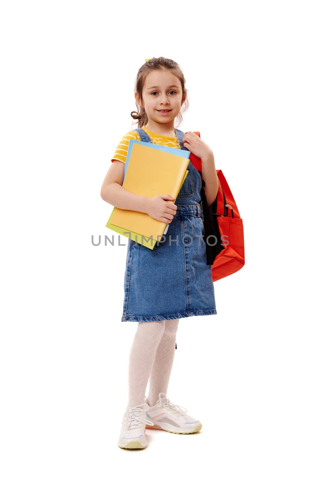 Full length portrait of Caucasian beautiful little girl, school kid holding backpack and studying books, smiling and looking confidently at camera, isolated on white background. Back to school concept