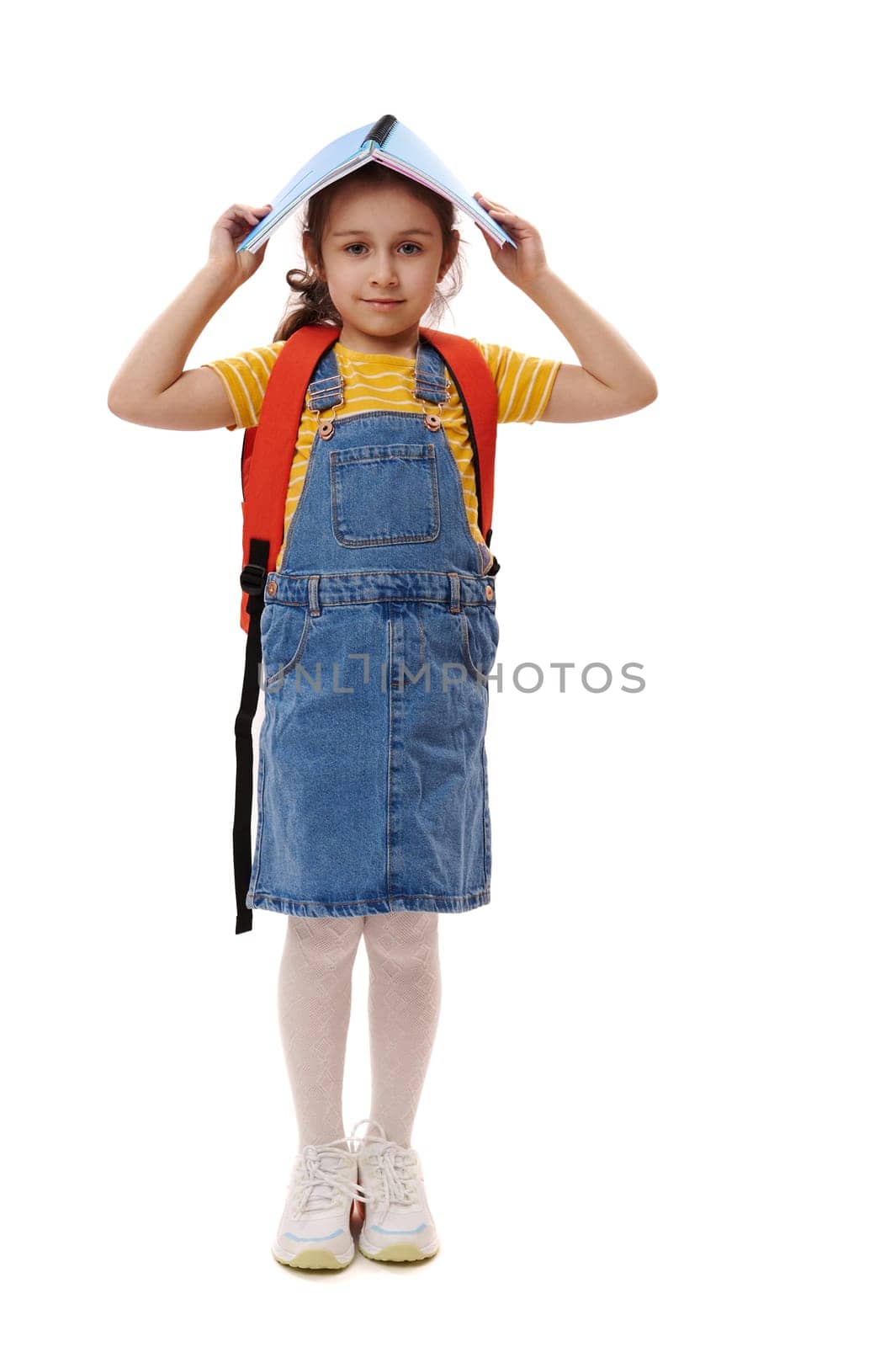 Full length smart student child kid,positive pleasant first grader holding open book above her head, smiling cutely looking at camera, isolated over white background. Back to school. Education concept