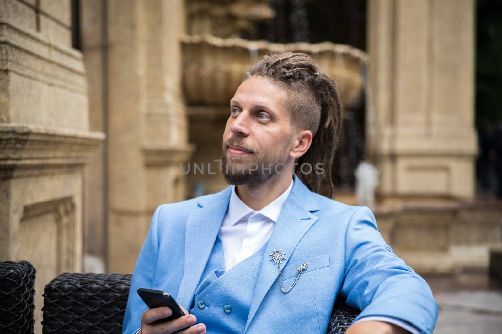 A young successful man in a blue suit with a smartphone in his hands sits in a luxury street restaurant.