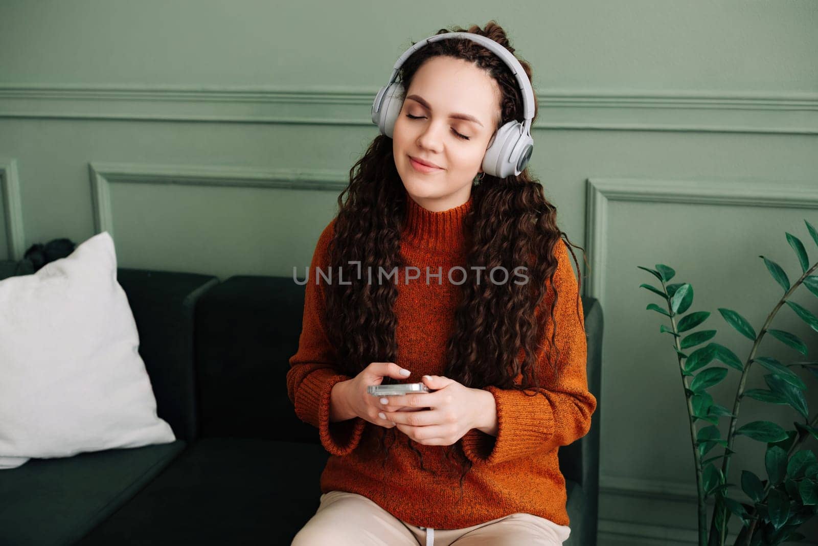 A calm, relaxed, carefree young woman listens to music, an audio podcast or audiobook on her cell phone while sitting at home, meditating and relaxing, feeling peace of mind with her eyes closed
