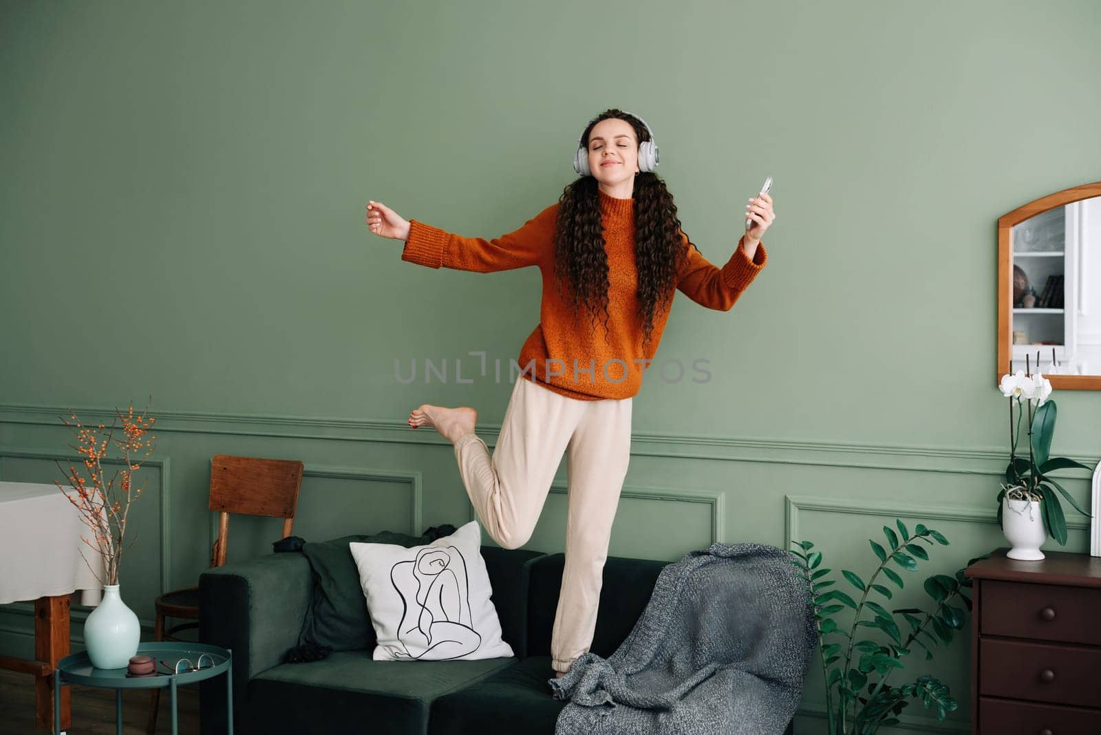 A happy, pretty young woman wearing headphones dances to her favorite song at home. Cheerful funny girl listening to popular music, moving, jumping on the couch in the living room, experiencing joy
