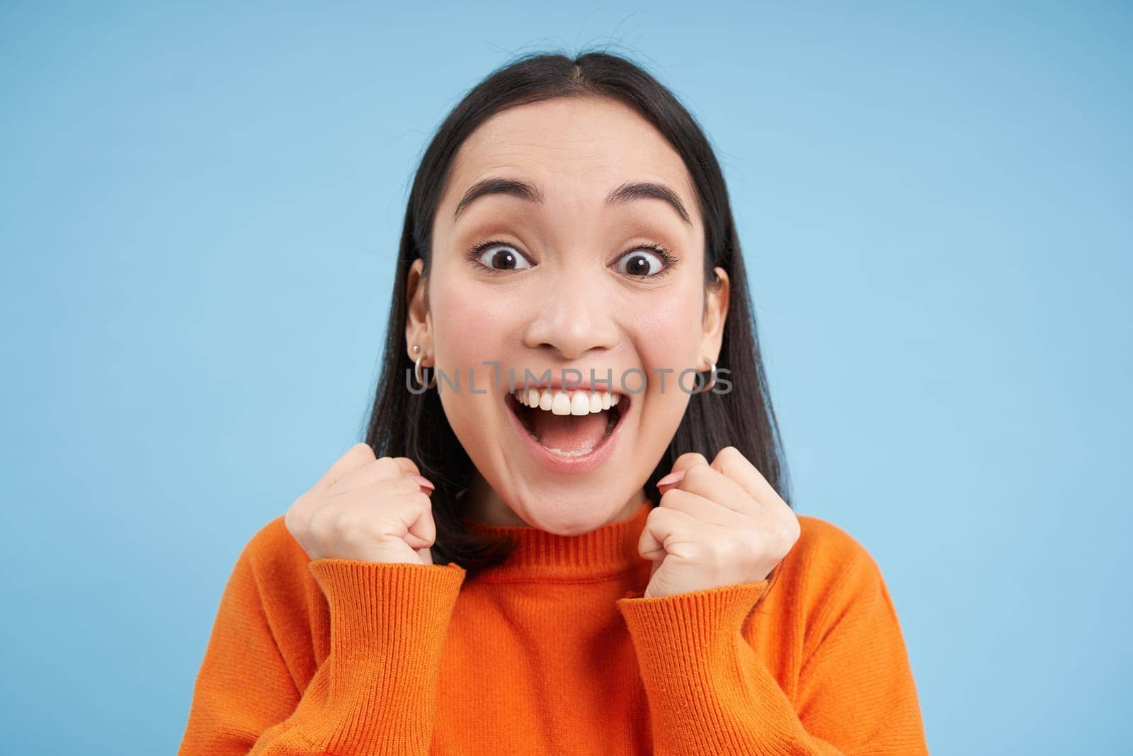 Close up of enthusiastic asian woman celebrates, wins and screams of joy, jumps from excitement, stands over blue background.